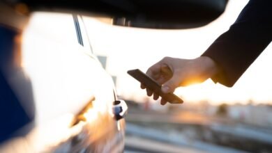 Person unlocking car door with smartphone, sunset background.