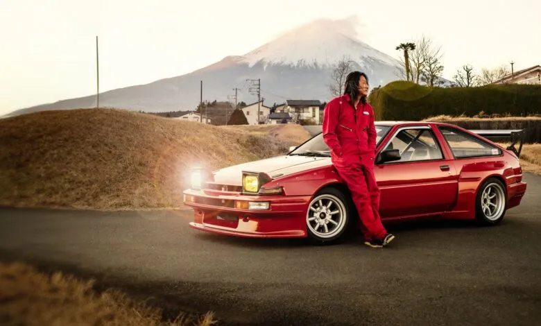 Sung Kang leans against a red Toyota AE86 with Mount Fuji in the background.