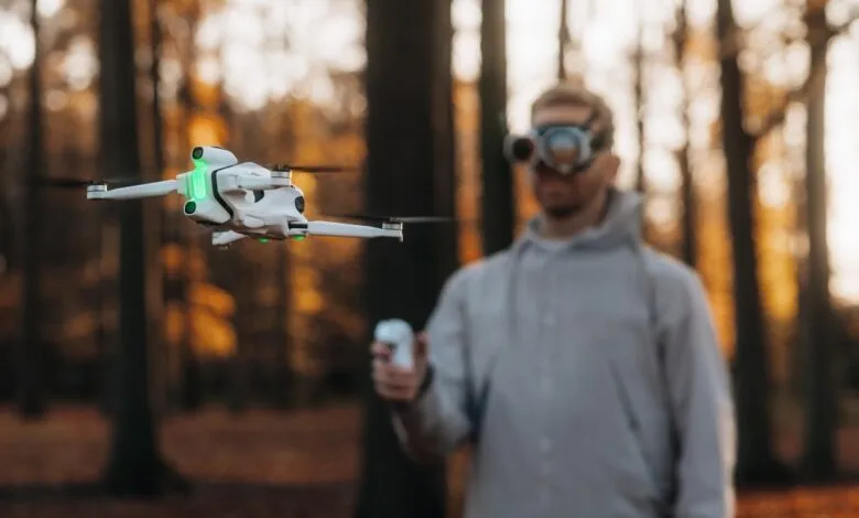 A white drone hovers in front of a blurred man wearing goggles in a forest.
