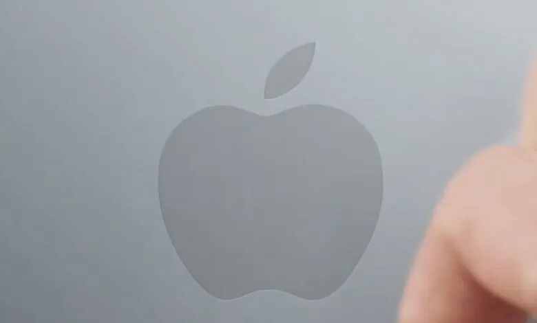 Apple logo on a gray surface with a blurred finger in the foreground.