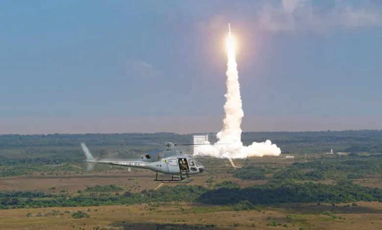 Helicopter flies as rocket launches, plume of smoke rises against blue sky.