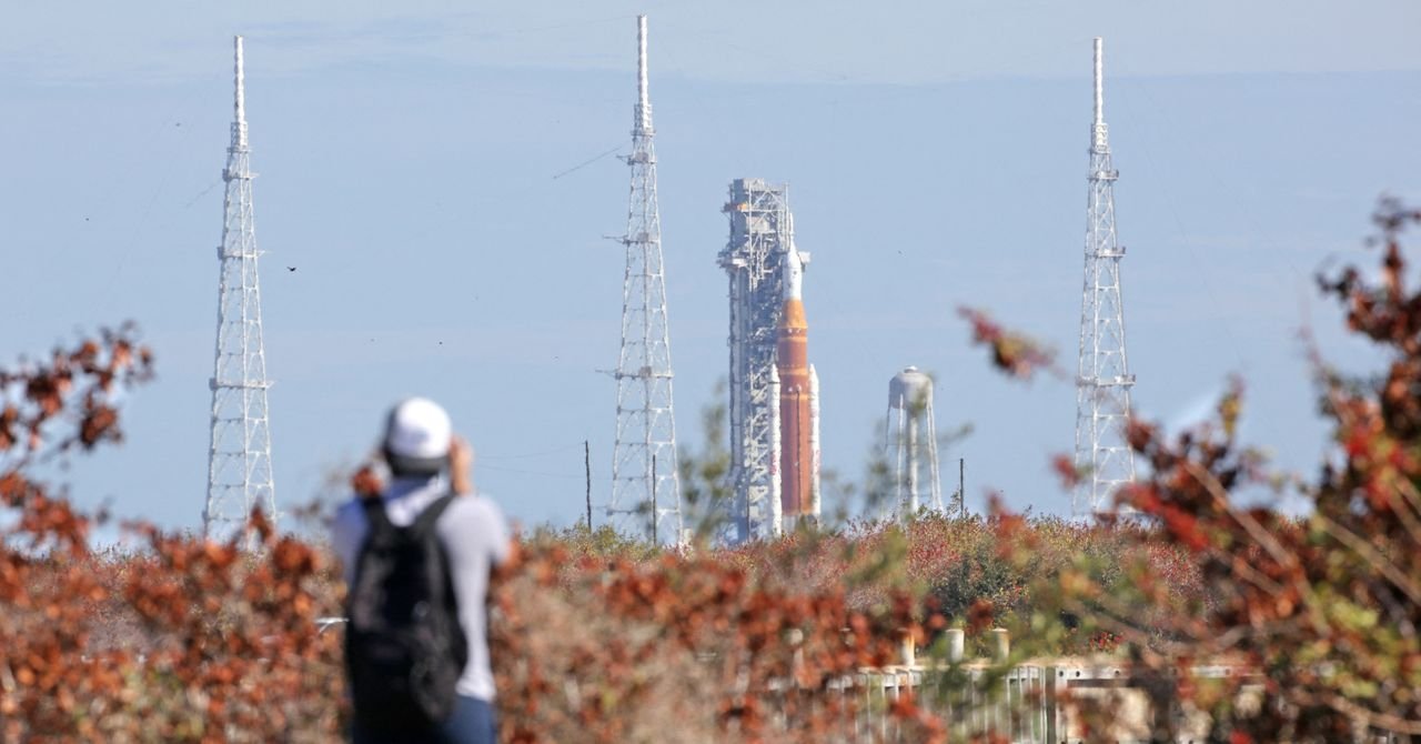 Artemis rocket on launchpad viewed through foliage and towers, person taking photo.
