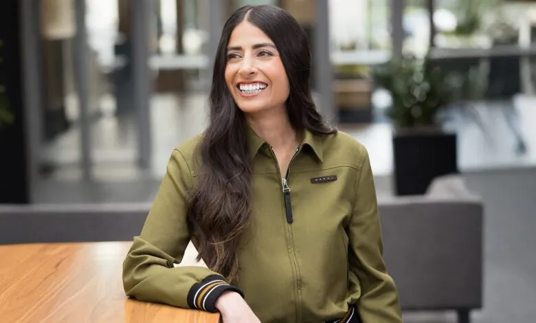 Asha Sharma smiles, wearing a green jacket and leaning on a wooden table.
