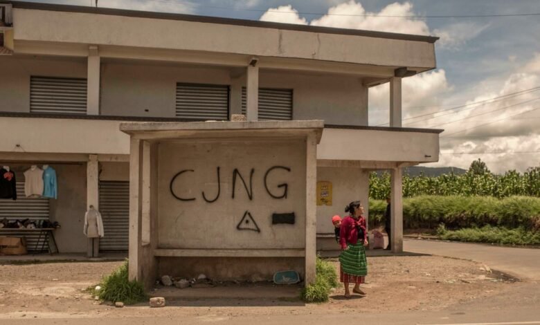 Woman with child stands near bus stop graffitied with 'CJNG'.