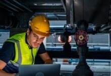 Technician in hard hat and vest using laptop near industrial equipment.