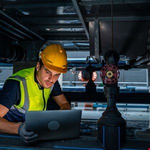 Technician in hard hat and vest using laptop near industrial equipment.