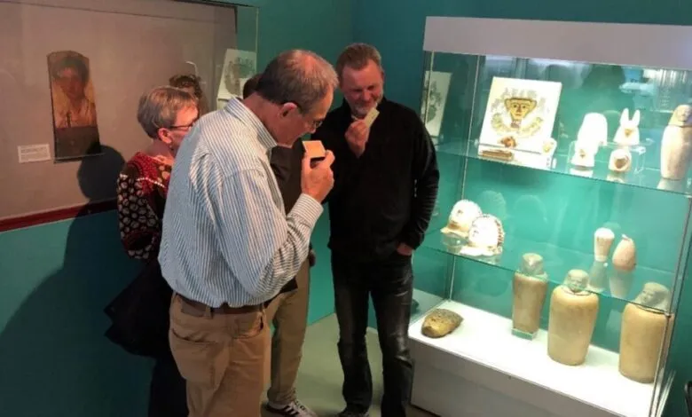Museum visitors smelling samples near Egyptian canopic jars in display case.