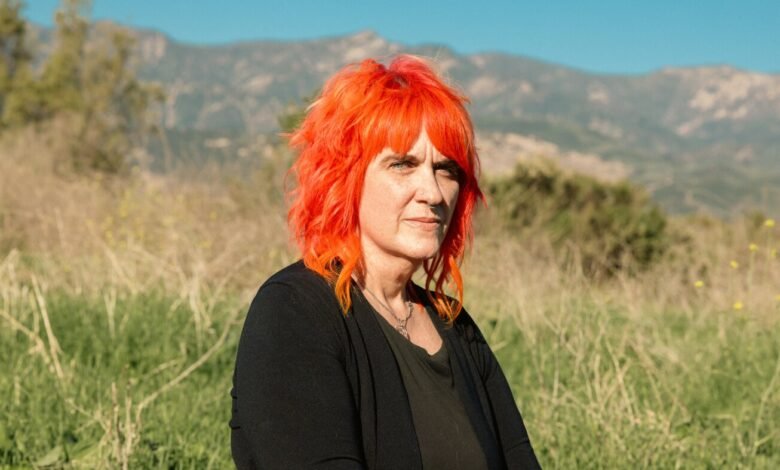Woman with bright orange hair sits in a field with mountains in the background.
