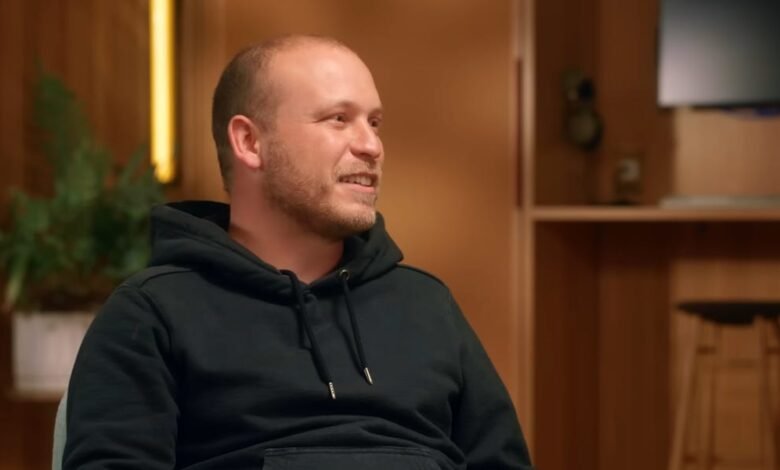 Man with short hair and black hoodie smiles in a warm, inviting room.