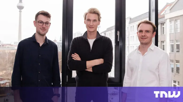 Three young men stand in front of a window with a city view.