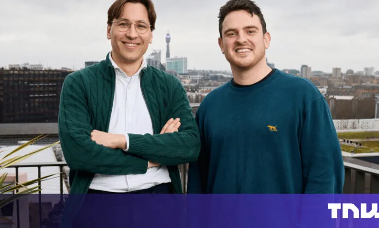 Two smiling men stand on a rooftop with a city skyline behind them.