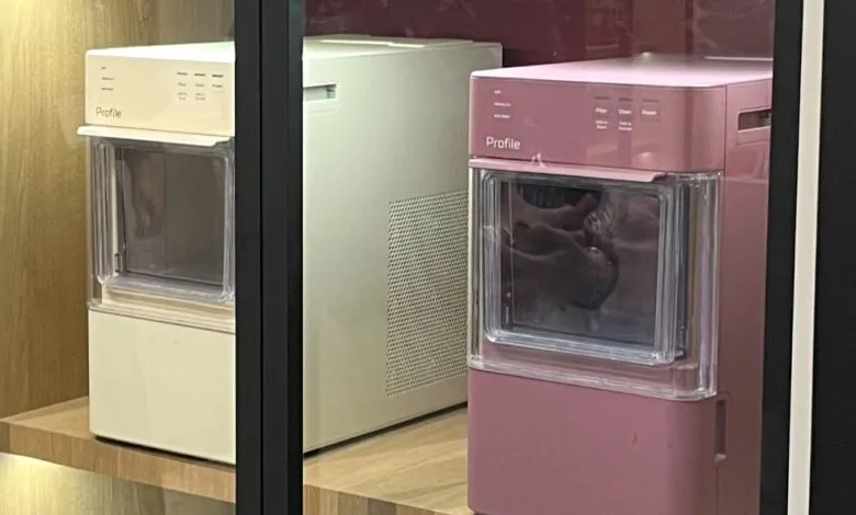 Two GE Profile ice makers, one cream and one pink, displayed on a wooden shelf.