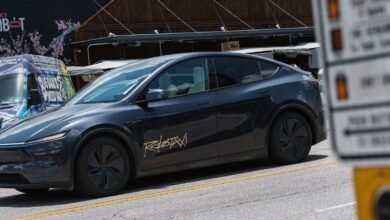 Dark gray Tesla Robotaxi parked on a street with a blurred sign in the foreground.