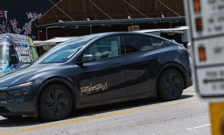 Dark gray Tesla Robotaxi parked on a street with a blurred sign in the foreground.
