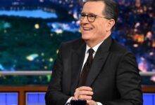 Stephen Colbert smiles on set in a dark suit and glasses.