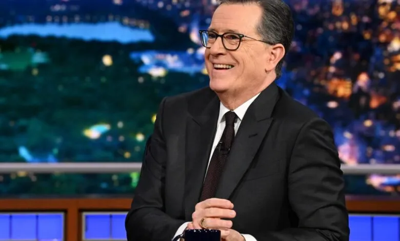 Stephen Colbert smiles on set in a dark suit and glasses.