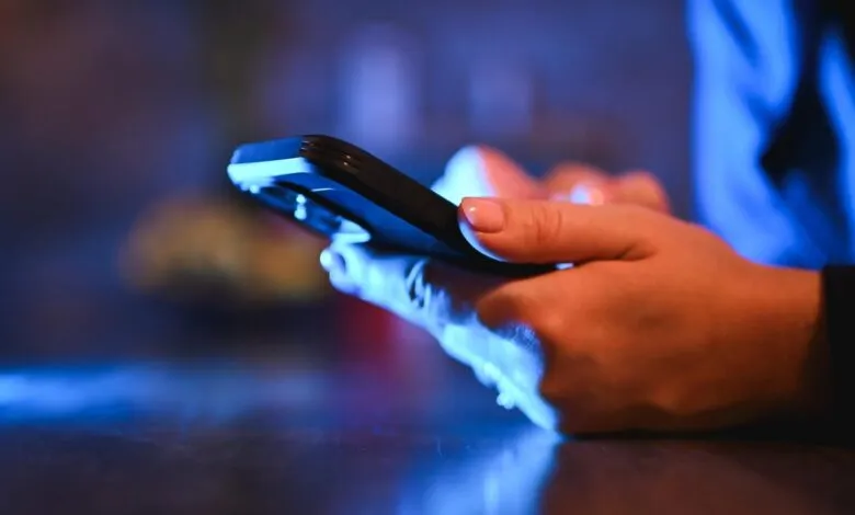 Close-up of hands using a smartphone in a dimly lit, blue-toned environment.