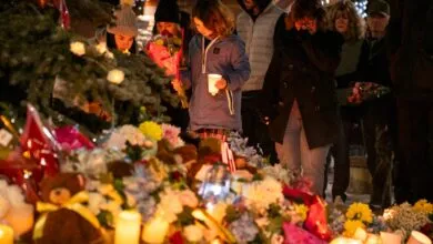 Mourners gather at memorial with flowers, candles, and teddy bears.