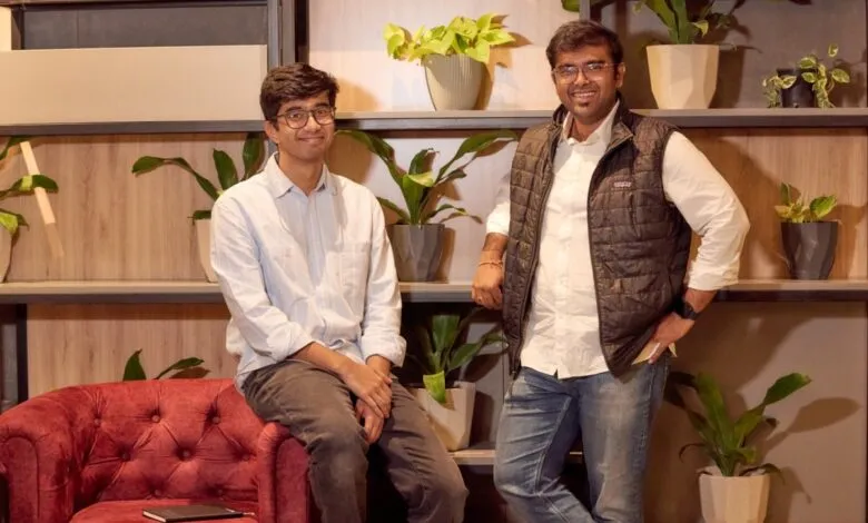 Two men pose in front of a shelf with plants, one seated on a red chair.