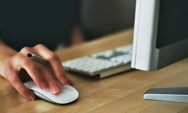 Hand holding stylus over computer mouse on wooden desk.
