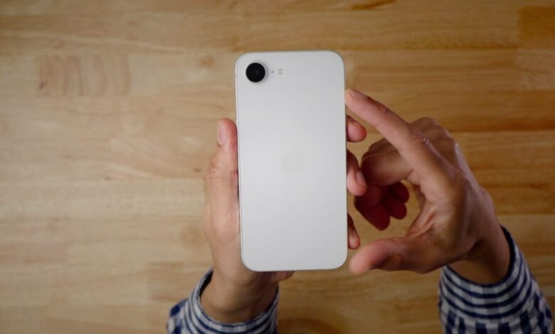 Hands holding a white iPhone with a single camera lens on a wooden surface.