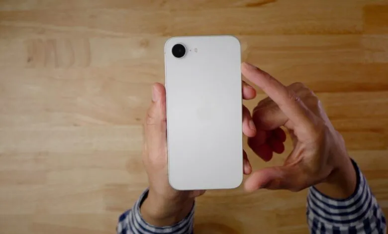 Hands holding a white iPhone with a single camera lens on a wooden surface.