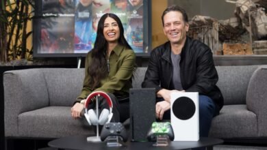 Sarah Bond and Phil Spencer smiling on a couch with Xbox consoles and accessories.