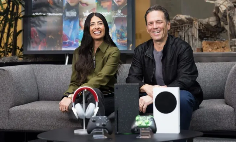 Sarah Bond and Phil Spencer smiling on a couch with Xbox consoles and accessories.