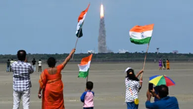 People wave Indian flags as a rocket launches into the sky, celebrating space exploration.