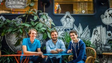 Three smiling men sit at a colorful table against a mural backdrop.