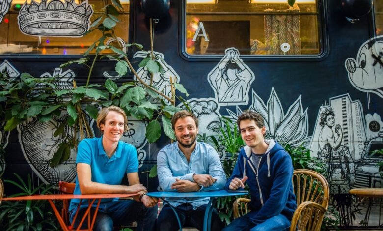 Three smiling men sit at a colorful table against a mural backdrop.