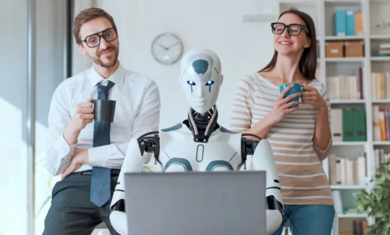 Robot working on laptop with two human colleagues holding coffee mugs.