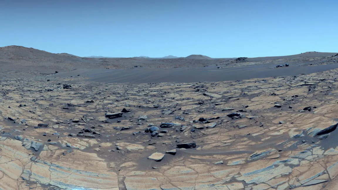 Panoramic view of the Martian surface with rocks, sand, and distant hills under a blue sky.