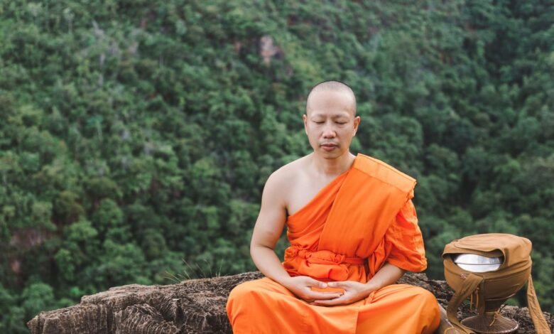 Monk in orange robe meditating on a rocky outcrop with forest backdrop.