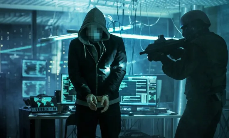 Hacker in handcuffs faces armed officer in a dark, tech-filled room.