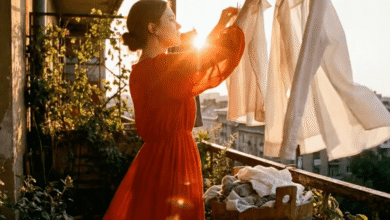 Woman in red dress hangs laundry on sunlit balcony with cityscape view.