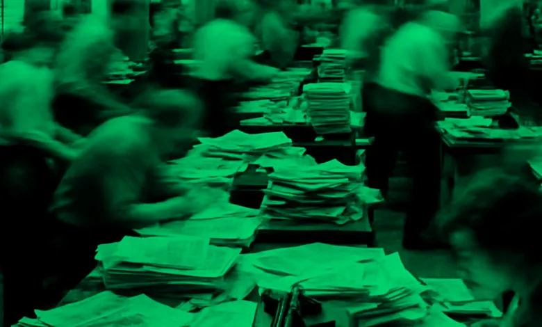 Blurred image of people working at desks covered in stacks of papers, green filter.