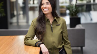 Smiling woman in green jacket leans on wooden table indoors.