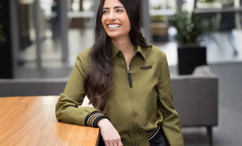 Smiling woman in green jacket leans on wooden table indoors.