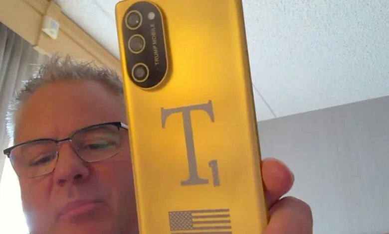 Man holds up a gold phone with 'Trump Mobile' and an American flag design.