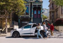 Three people stand near a white Waymo car in front of colorful San Francisco houses.