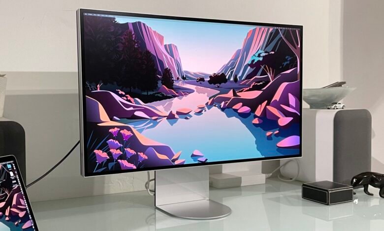 Studio Display showing landscape art, with laptop, speakers, and decorative objects.