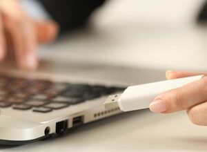 Person inserting a white USB drive into a silver laptop on a white desk.