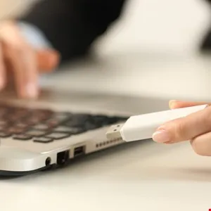 Person inserting a white USB drive into a silver laptop on a white desk.