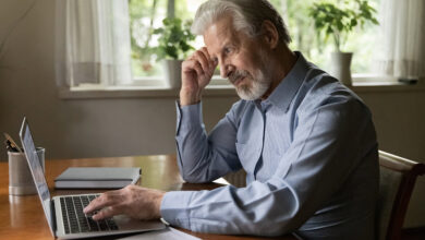 Senior man thoughtfully using laptop at desk in a bright room.