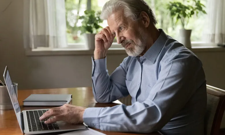 Senior man thoughtfully using laptop at desk in a bright room.