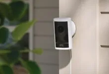 Ring security camera mounted on a light beige exterior wall with green foliage nearby.