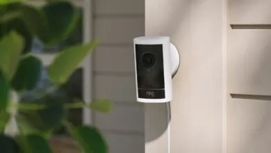Ring security camera mounted on a light beige exterior wall with green foliage nearby.