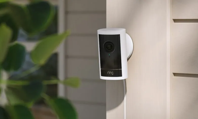 Ring security camera mounted on a light beige exterior wall with green foliage nearby.