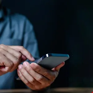 Close-up of hands using a smartphone on a wooden surface against a dark background.